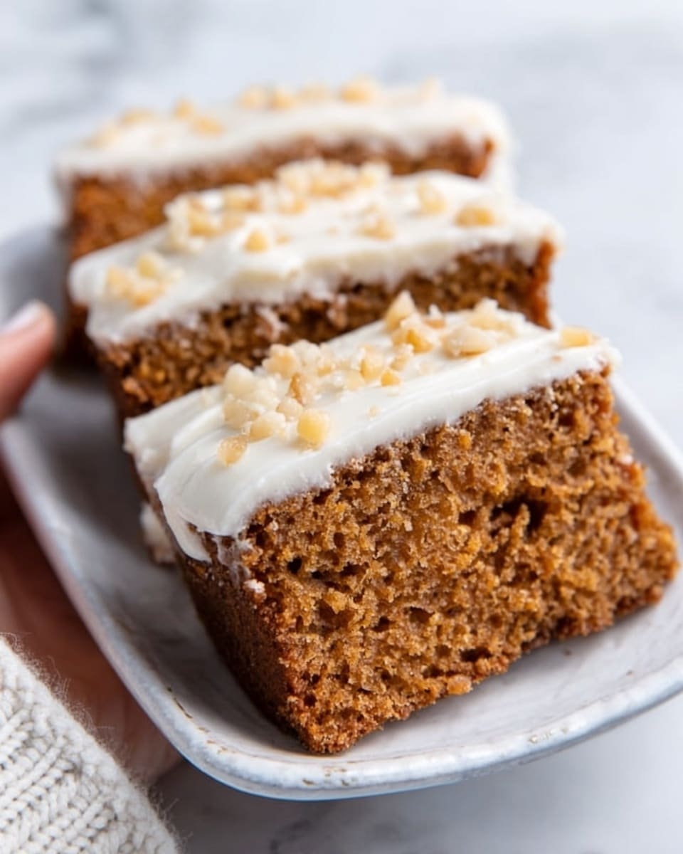 A close-up of a two-layer rectangular slice of brown cake on a white plate, sitting on a white marbled surface. The cake has a soft, slightly crumbly texture with a thick layer of smooth white icing spread evenly on top, decorated with small crushed nuts scattered across the icing. A woman's hand is gently holding the edge of the plate on the side. Photo taken with an iphone --ar 4:5 --v 7